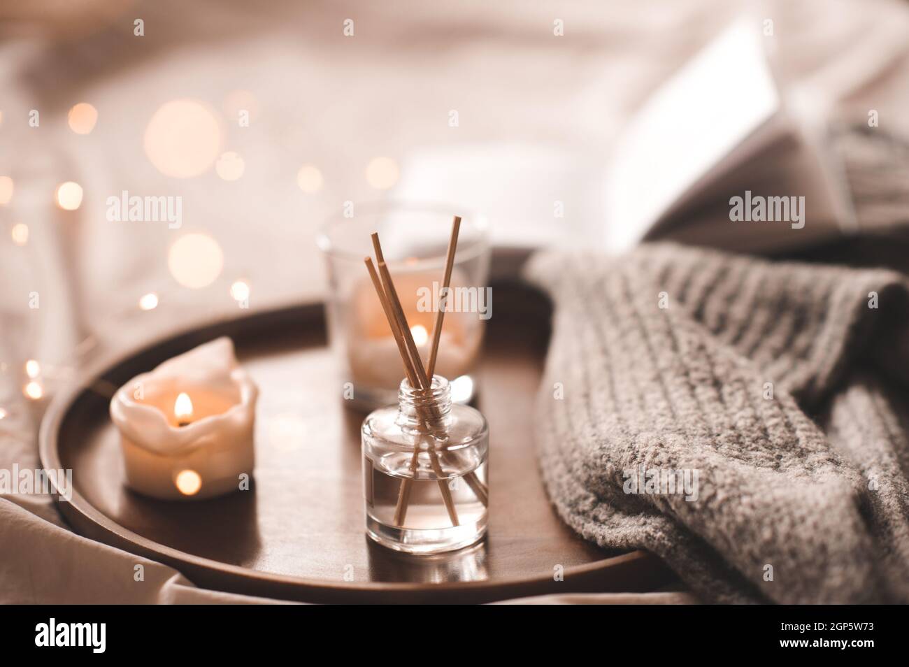 Profumo domestico in bottiglia di vetro con i bastoni di legno, candele profumate di bruciatura, libro aperto di carta e tessuto di lana di maglia sul vassoio in primo piano della camera da letto. Aromaterapia c Foto Stock