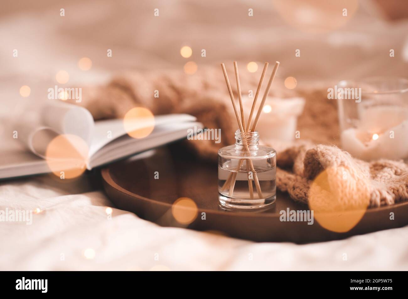 Profumo domestico in bottiglia di vetro con i bastoni di legno, candele profumate di bruciatura, libro aperto di carta e tessuto di lana di maglia sul vassoio in primo piano della camera da letto. Aromaterapia c Foto Stock