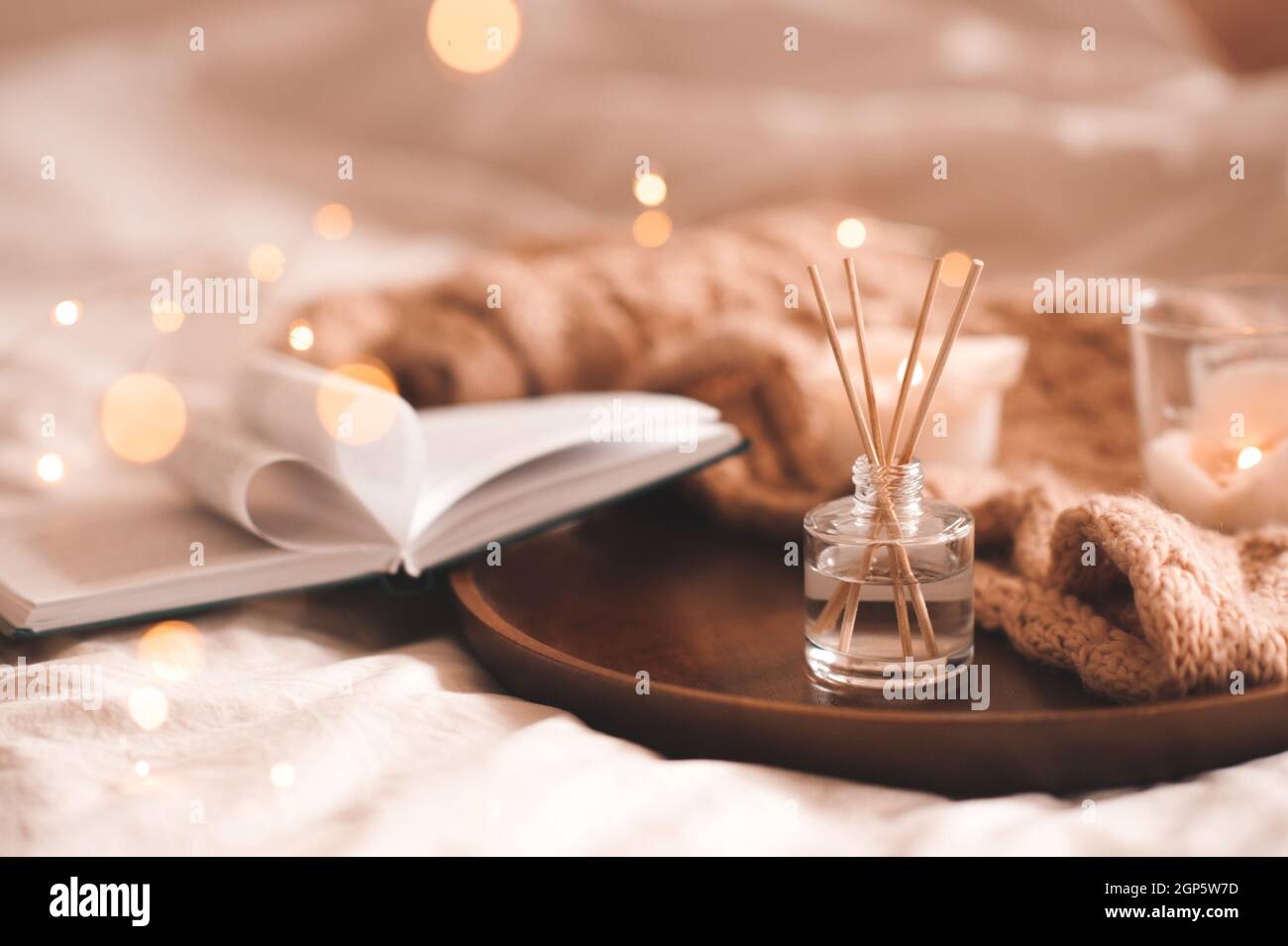 Profumo domestico in bottiglia di vetro con i bastoni di legno, candele profumate di bruciatura, libro aperto di carta e tessuto di lana di maglia sul vassoio in primo piano della camera da letto. Aromaterapia c Foto Stock