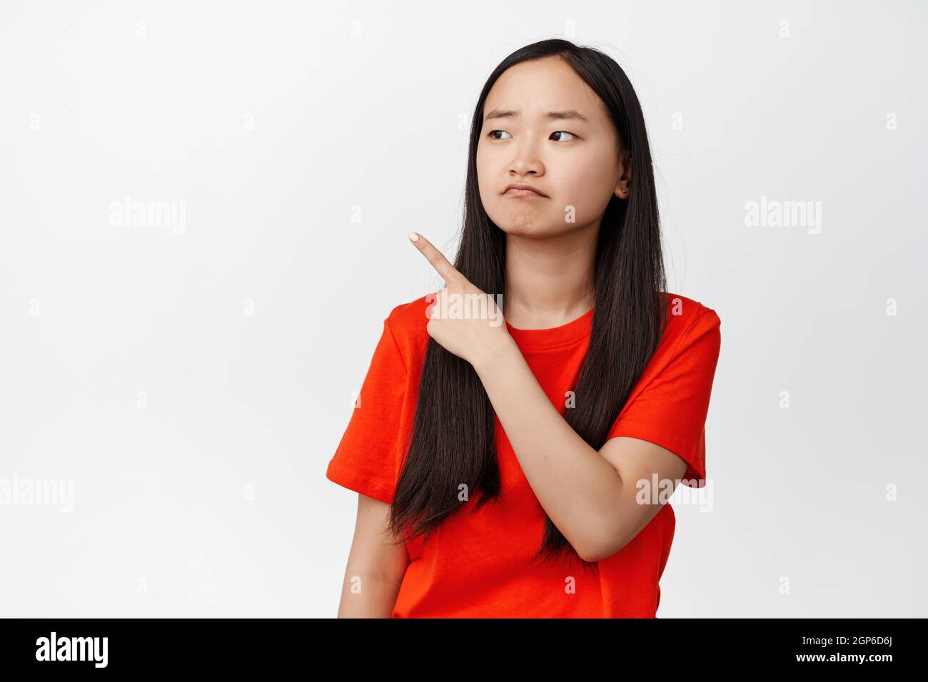 Deluso ragazza asiatica teen mostra rammarico, puntando e guardando a sinistra con accovacciamento volto, in piedi sconvolto su sfondo bianco Foto Stock