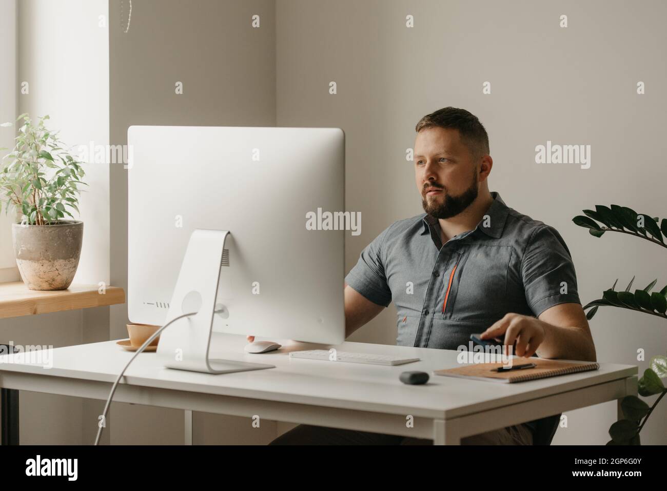 Un uomo lavora in remoto su un computer desktop. Un ragazzo con una barba sta mettendo un telefono cellulare giù durante un rapporto di un collega ad una videoconferenza a ho Foto Stock