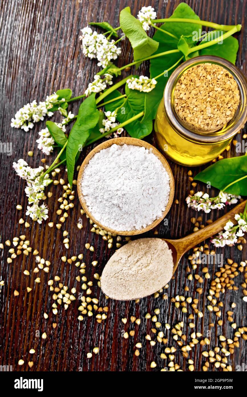 Farina di grano saraceno da cereali verdi in un recipiente, farina di grano saraceno da semole marroni in un cucchiaio, fiori e foglie sullo sfondo di un cinghiale di legno scuro Foto Stock