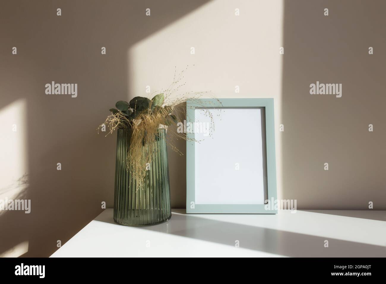Vaso di vetro con fiori di erbario, rami di eucalipto, cornici bianche vuote sul tavolo bianco all'interno con pareti beige vicino alla finestra. Foto Stock
