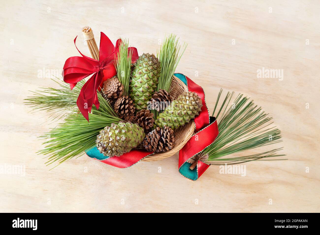 Coni di pino, verdi e marroni, in un cestino di vimini su sfondo di legno chiaro. Ribon rosso e arco. Spazio copia. Foto Stock