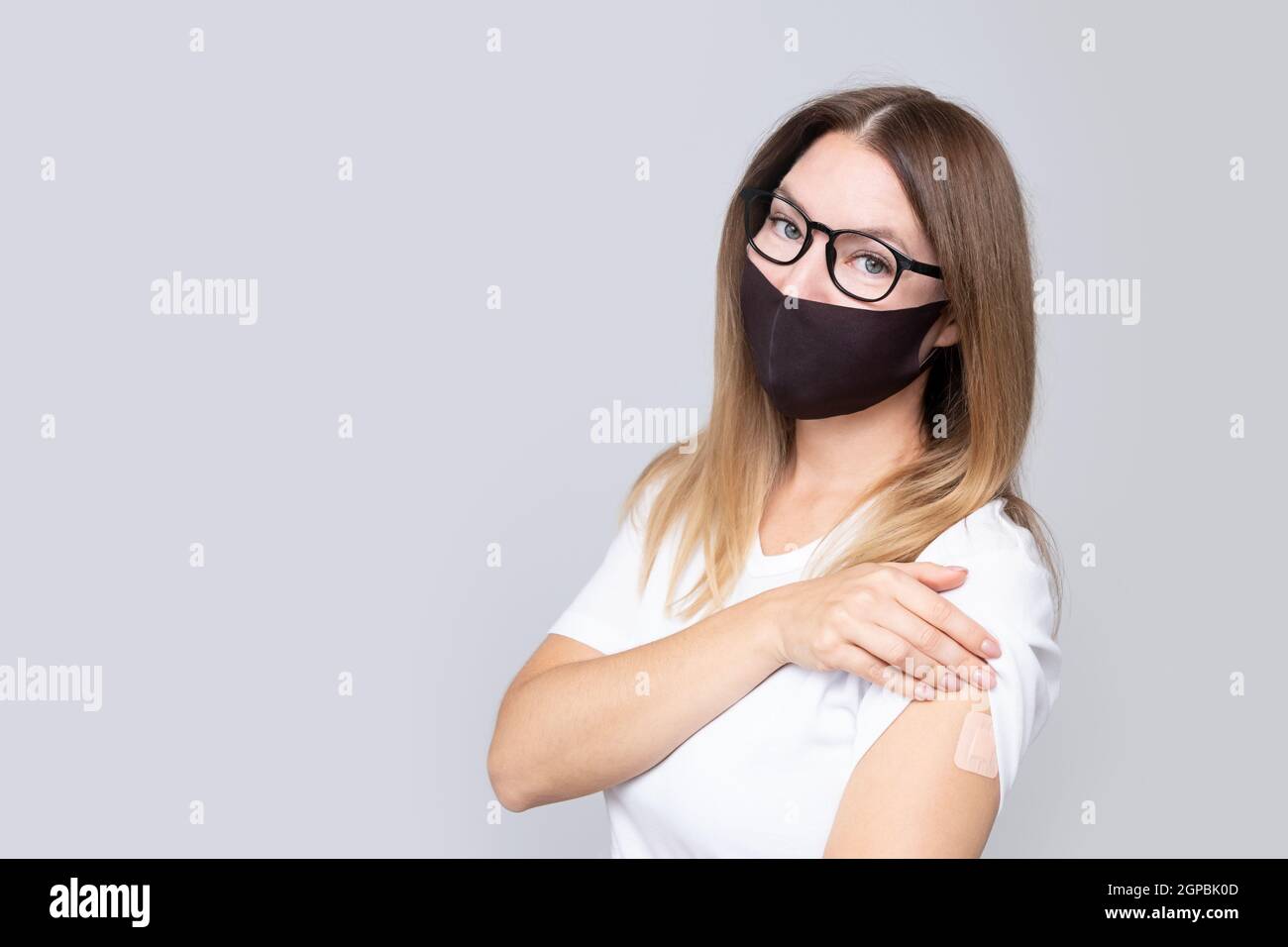 Giovane sorridente donna in vera mezza età in maschera facciale e T-shirt bianca e occhiali con un gesso sulla mano gioisce di ricevere una vaccinazione contro Foto Stock