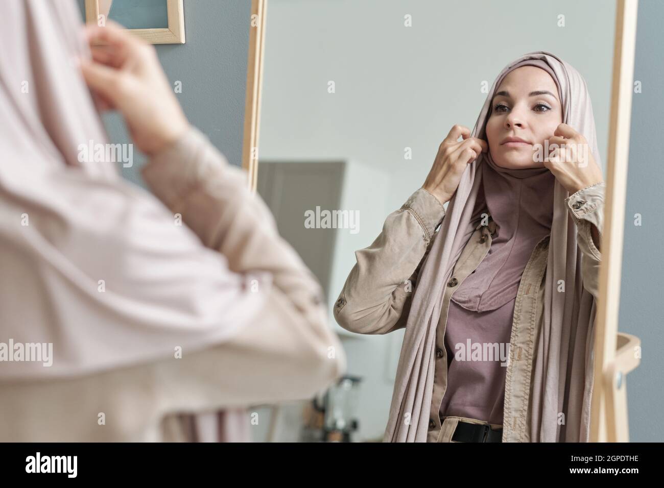 Scatto orizzontale sopra la spalla di attraente giovane donna musulmana adulta che si prepara ad uscire mettendo su hijab davanti allo specchio Foto Stock
