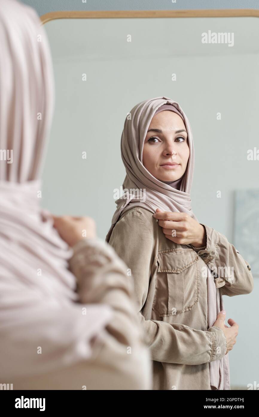 Colpo verticale sopra la spalla della giovane donna musulmana moderna adulta che si prepara ad uscire fissando hijab davanti allo specchio Foto Stock
