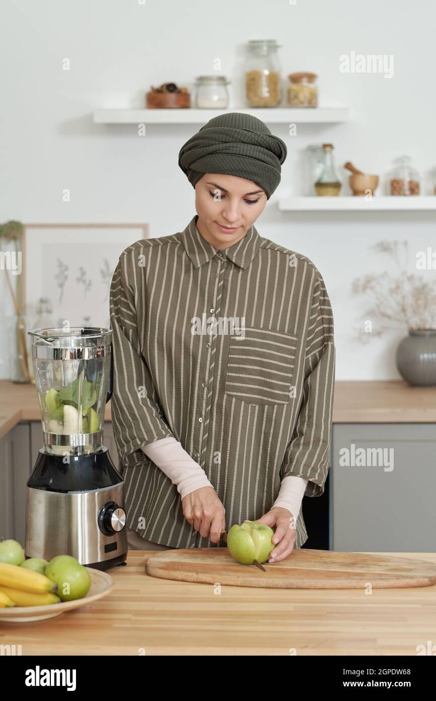 Verticale medio ritratto di bella donna musulmana vestito alla moda passare mattina in cucina taglio mela verde per frullato Foto Stock