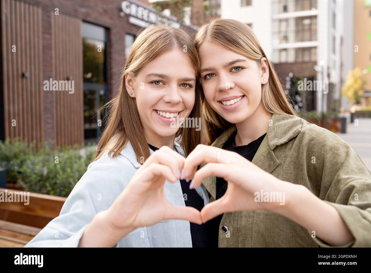 Buone ragazze gemelle affettuose in casualwear facendo la forma del cuore con le mani Foto Stock