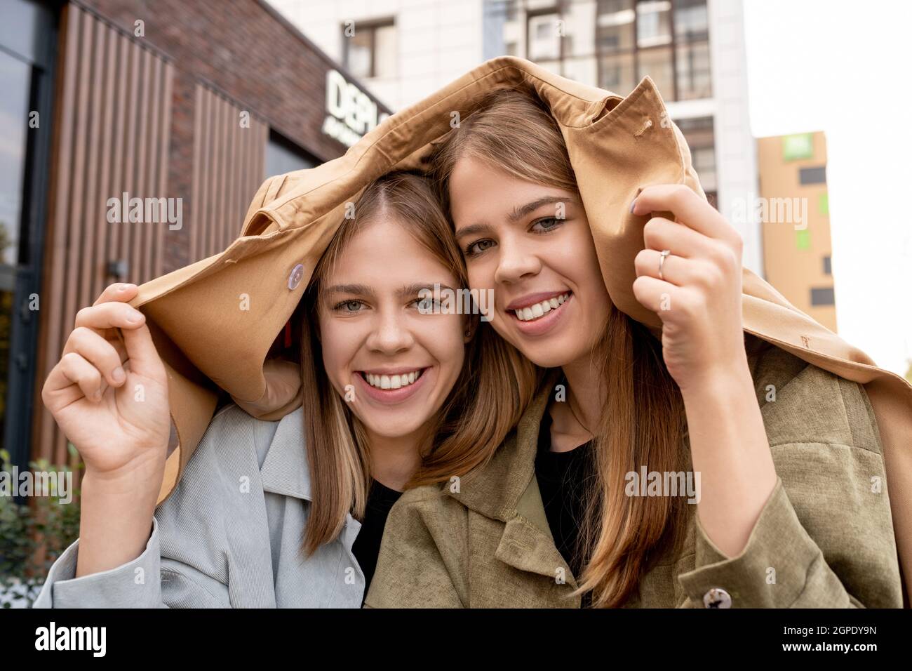 Due ragazze bionda felici che coprono le loro teste con trenchcoat beige in tempo piovoso Foto Stock