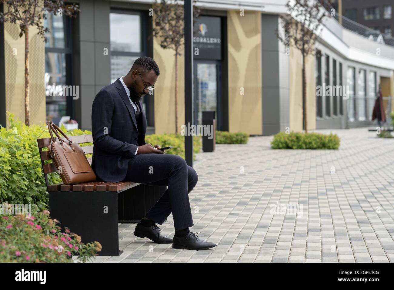 Giovane uomo d'affari africano che scorre in smartphone mentre si siede in panchina Foto Stock