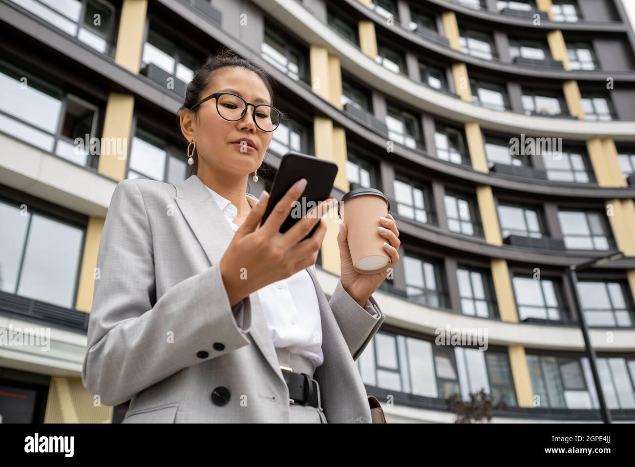 Giovane agente contemporaneo in abito elegante guardando lo schermo dello smartphone contro l'architettura moderna Foto Stock