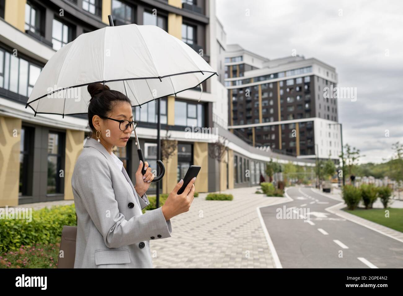 Giovane ed elegante donna d'affari che comunica in video chat sotto ombrello il giorno piovoso Foto Stock