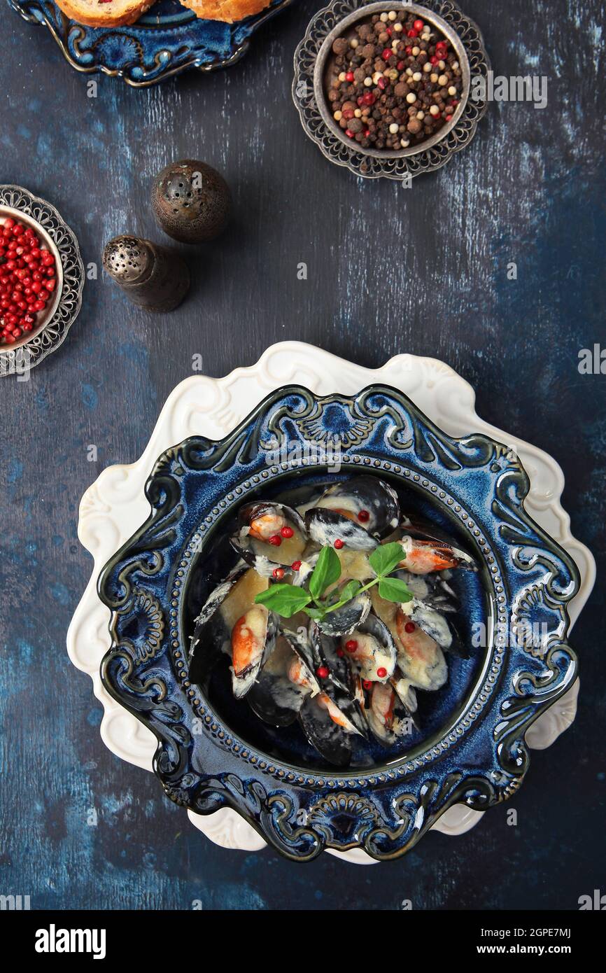 Cozze in salsa di panna. Frutti di mare su un piatto. Peperoni rossi e neri. Elegante vasellame in ceramica blu. Vista dall'alto. Sfondo blu. Spazio libero. Verticale Foto Stock