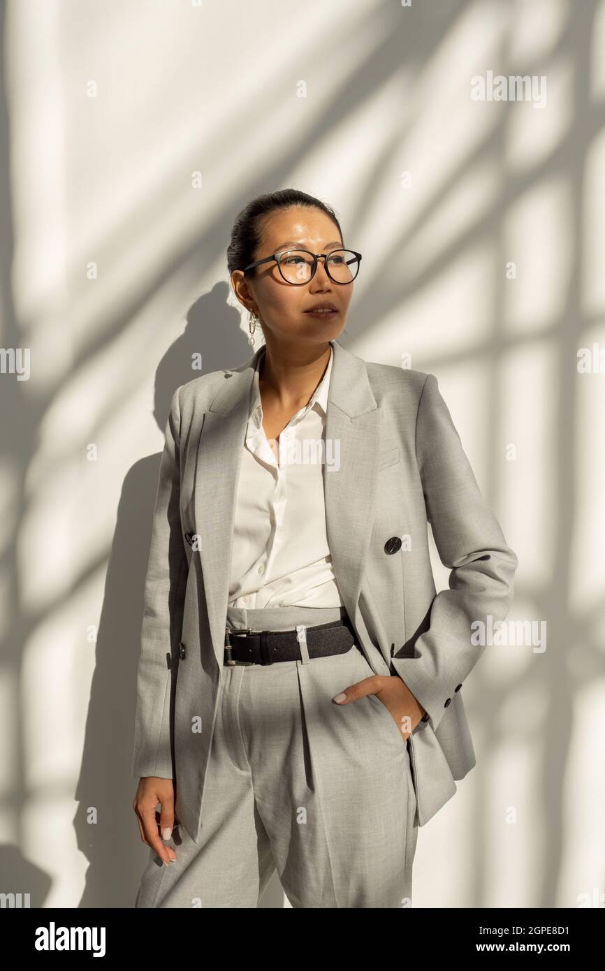 Donna d'affari asiatica contemporanea in elegante abito grigio che si erge contro il muro bianco Foto Stock