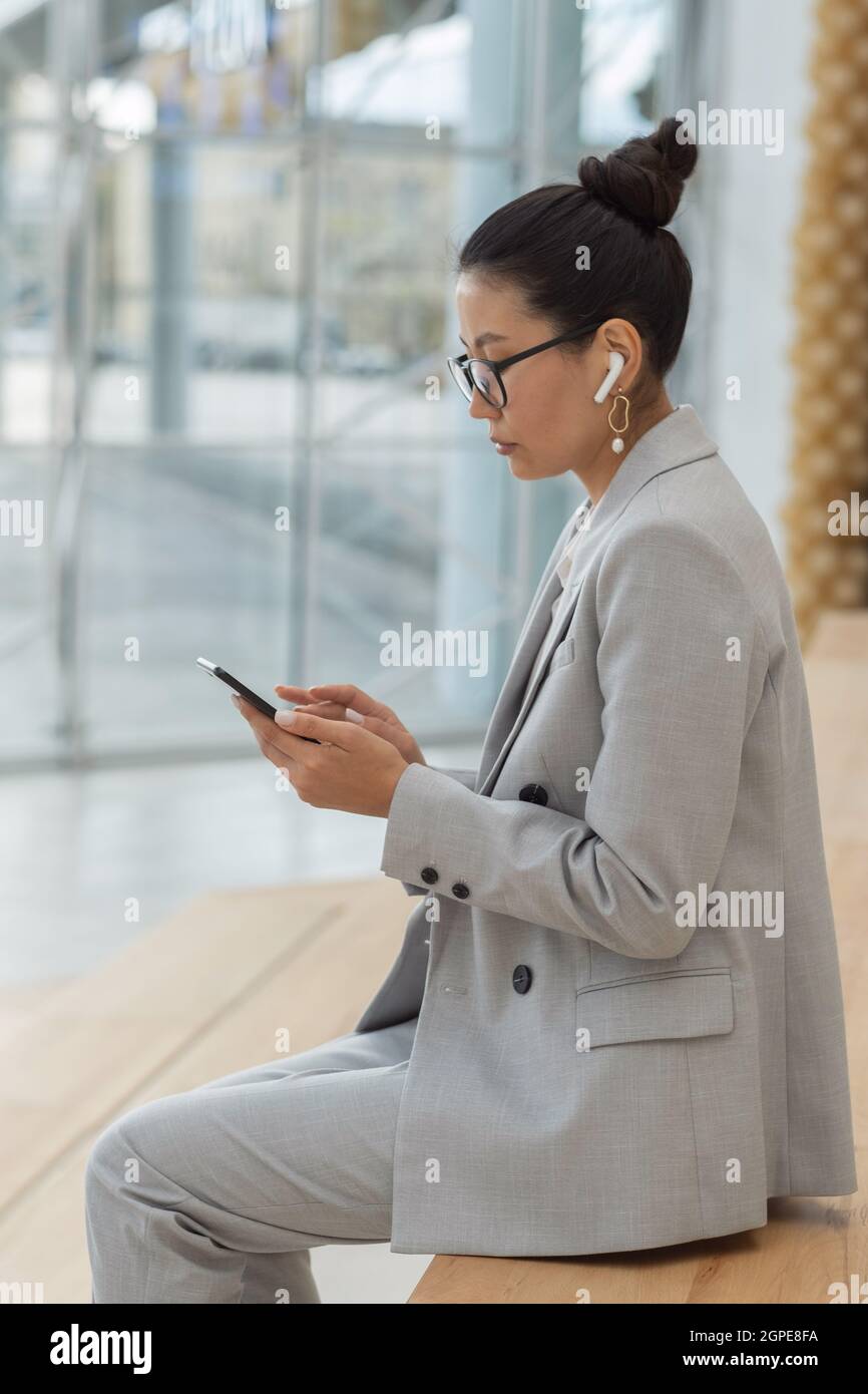 Giovane donna asiatica seria in abito, occhiali e cuffie guardando le notizie online smartphone Foto Stock
