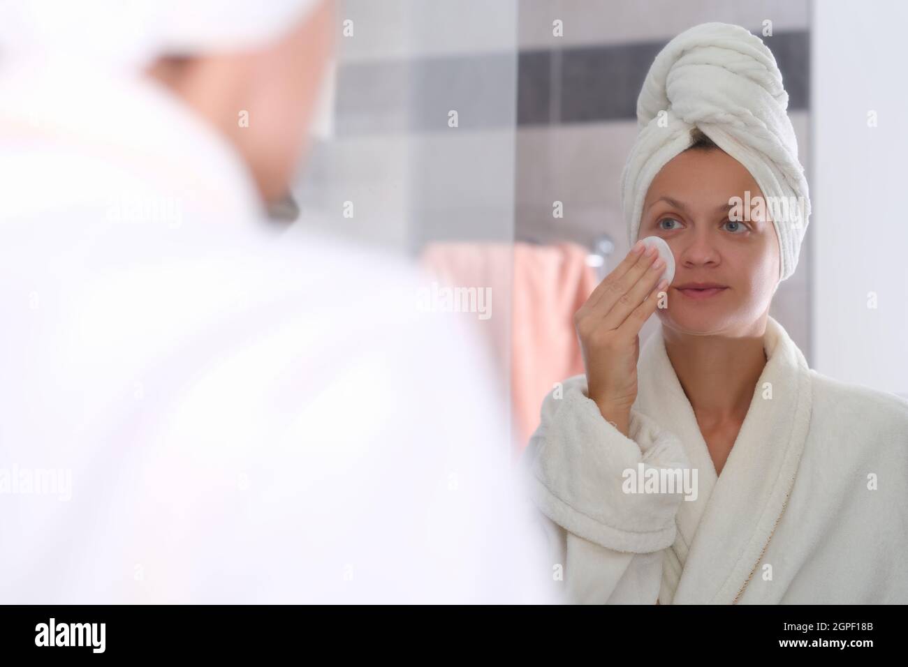 Giovane donna in accappatoio e asciugamano sulla testa lavare cosmetici dal viso di fronte allo specchio Foto Stock