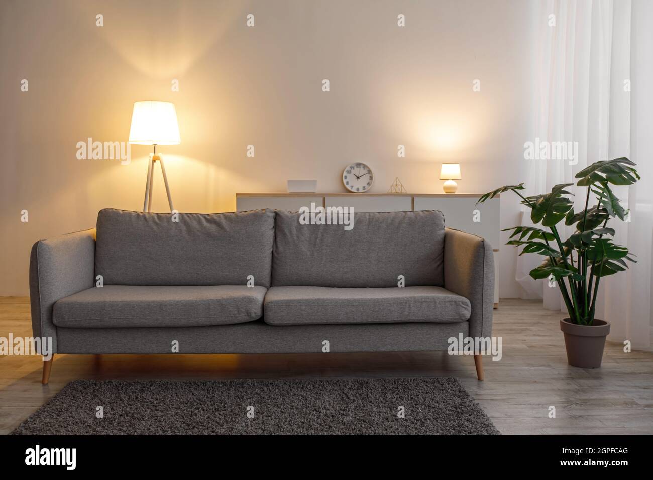 Divano retrò, tappeto, lampade incandescenti in serata, pianta in pentola sul pavimento e mobili in soggiorno interno Foto Stock