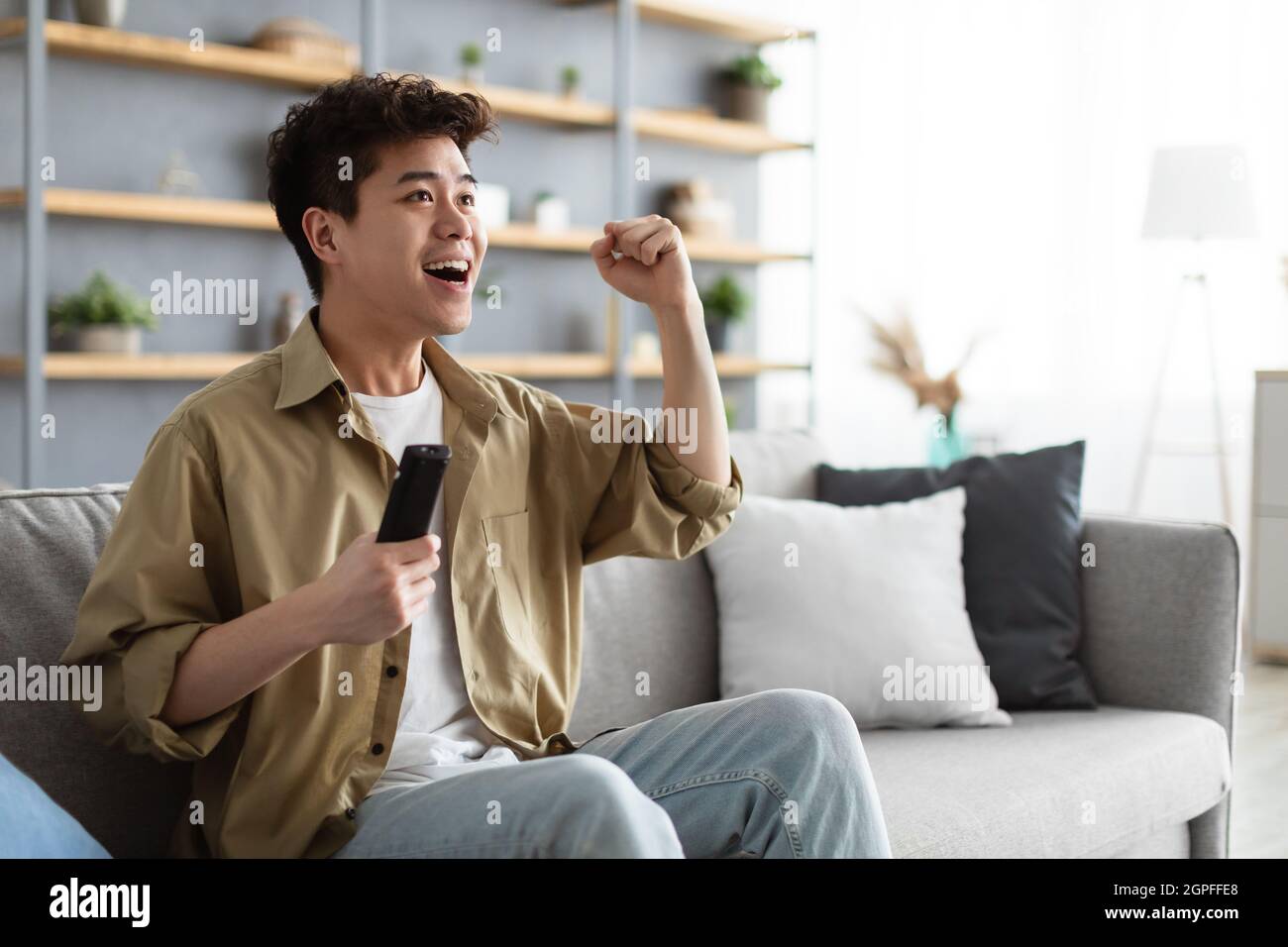 Felice ragazzo asiatico trascorrere il fine settimana guardando la tv scuotendo pugni clenched Foto Stock