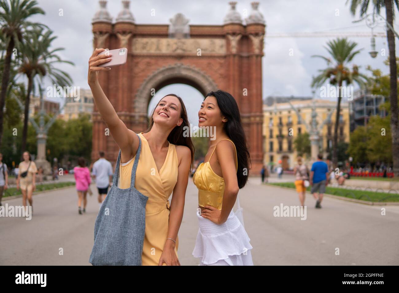 Felice giovane donna multirazziale amici in abiti eleganti che prendono selfie su smartphone durante il tour panoramico della città di Barcellona con arco trionfale sullo sfondo Foto Stock