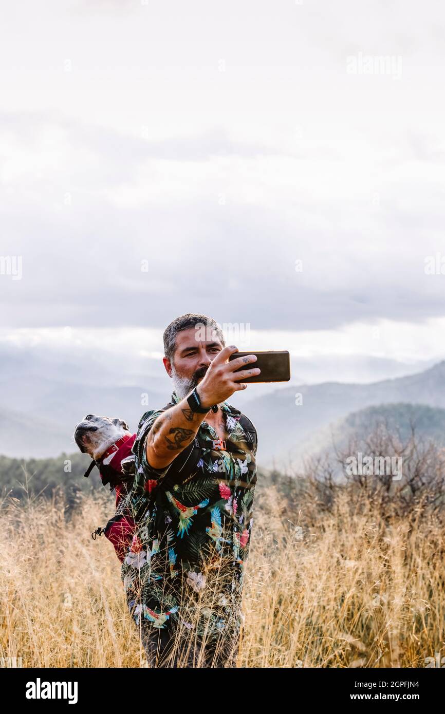 uomo con cane in zaino scattando foto sulla montagna con cielo nuvoloso Foto Stock