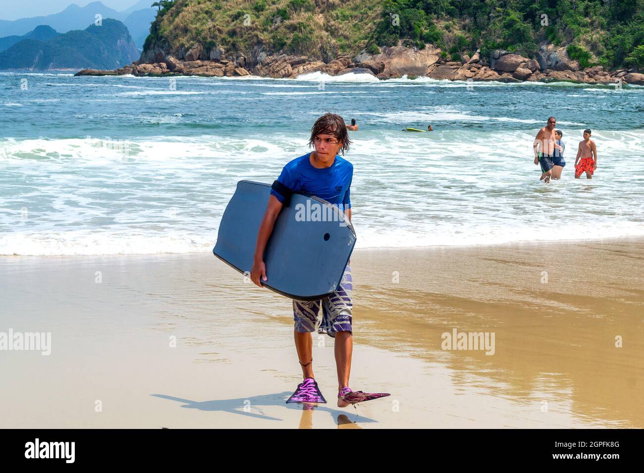 Giovane brasiliano con una tavola da surf sulla spiaggia Piratininga a Nitreoi, Rio de Janeiro, Brasile. Il punto di riferimento della spiaggia è un luogo famoso e una trave Foto Stock