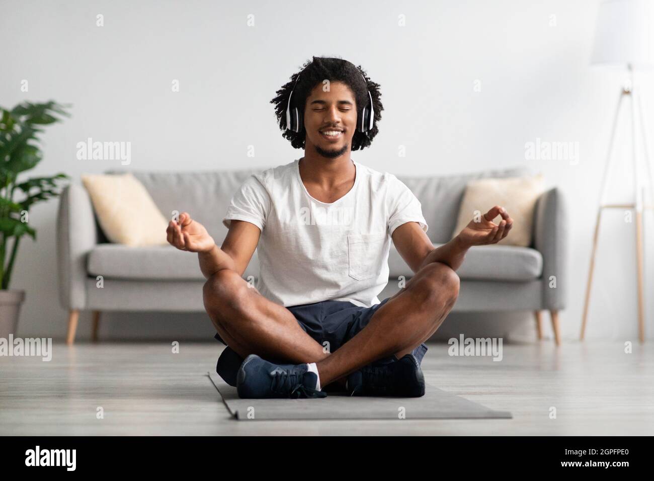 Concetto di meditazione. Calmo il ragazzo nero nelle cuffie wireless meditando a casa Foto Stock