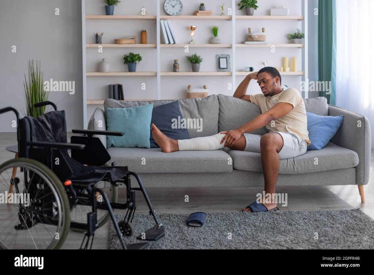Uomo nero infelice che ha bendato la gamba rotta, seduto sul divano, che soffre di dolore a casa, sedia a rotelle in piedi nelle vicinanze Foto Stock