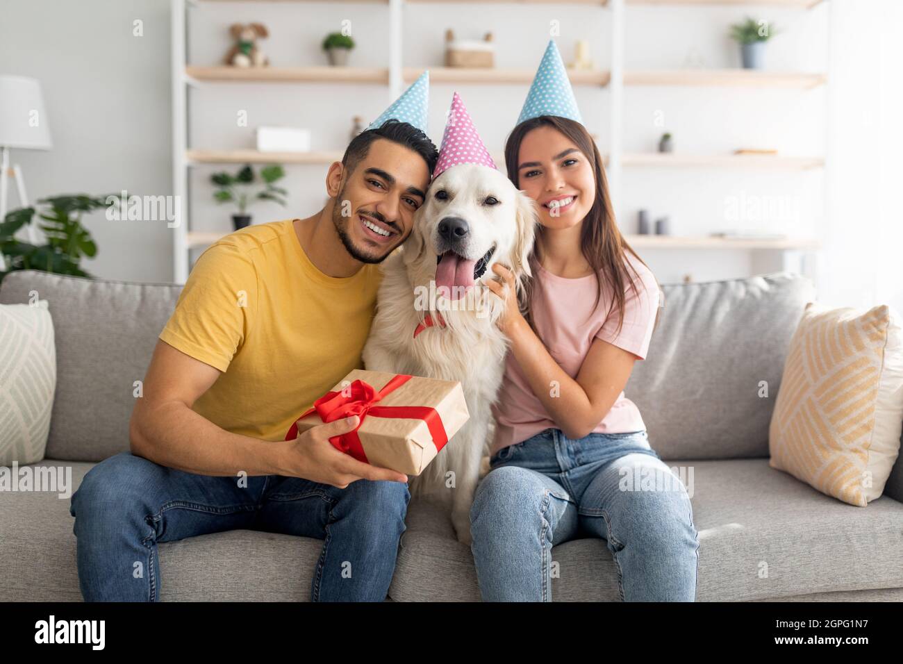 Felice coppia variegata celebrare il compleanno del loro cane del animale domestico, sedendosi sul divano con la scatola del regalo, portando i cappelli di festa nel paese Foto Stock