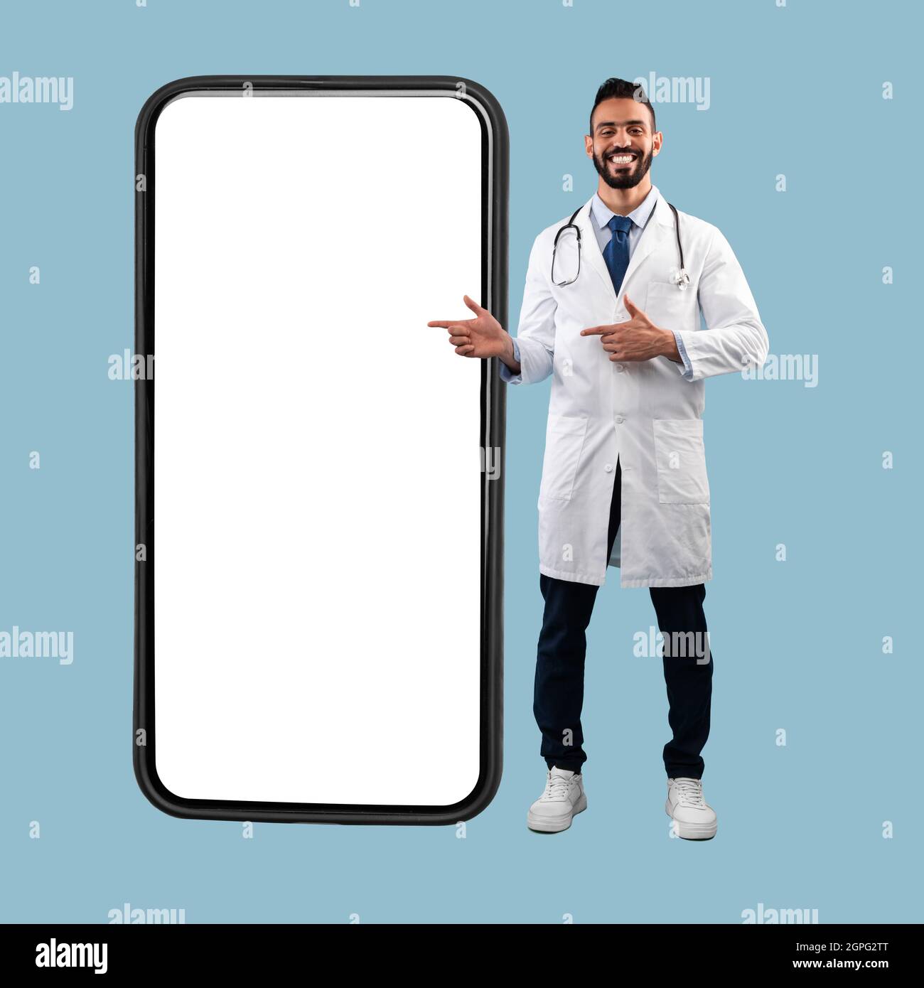 Medico medio-orientale che punta le dita sullo schermo del telefono grande, sfondo blu Foto Stock