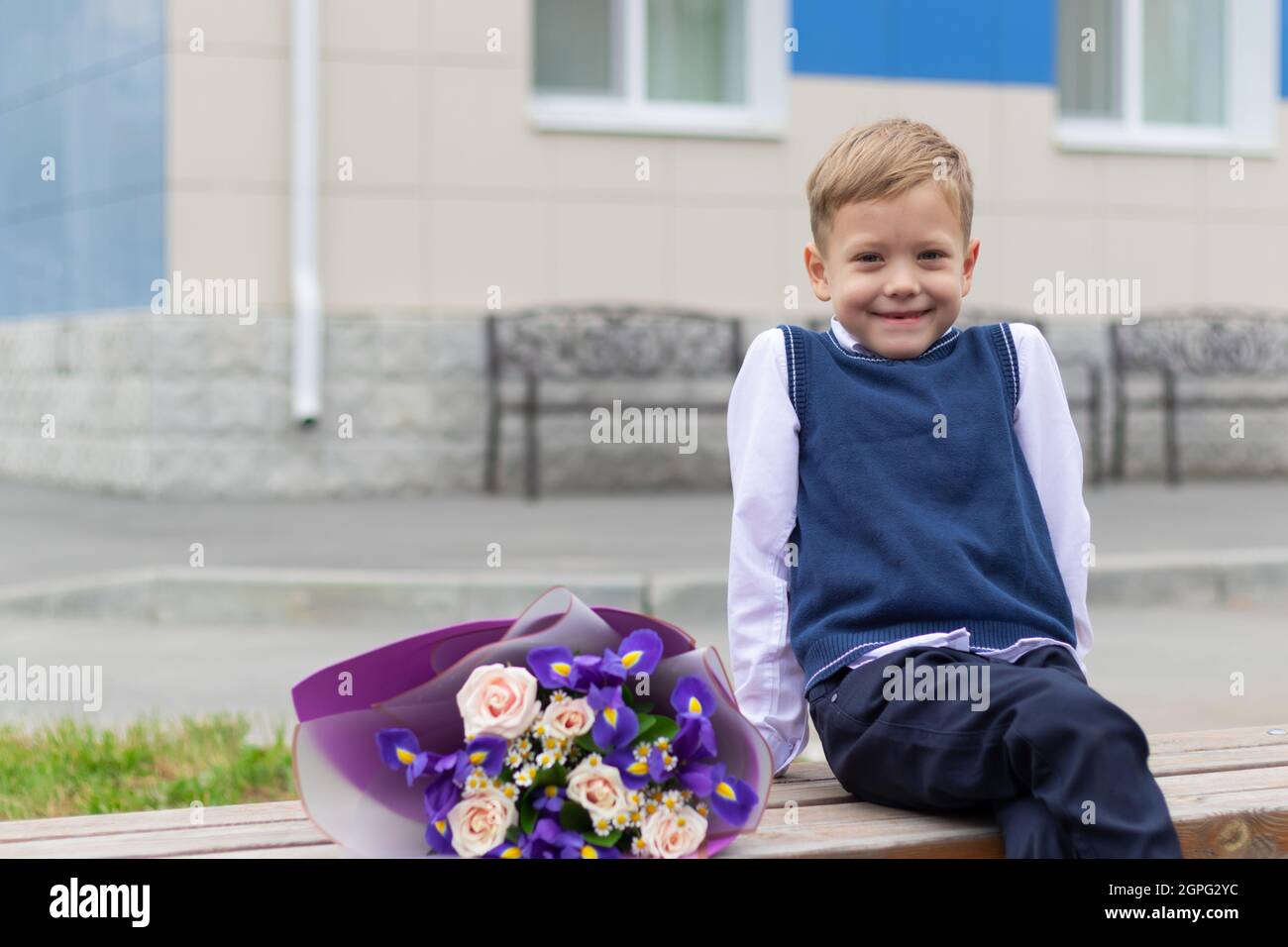 Un ragazzo carino primo-grader in un uniforme della scuola con un bouquet bello dei fiori colorati nel cortile della scuola dopo la festa il 1 settembre. Informazioni Foto Stock