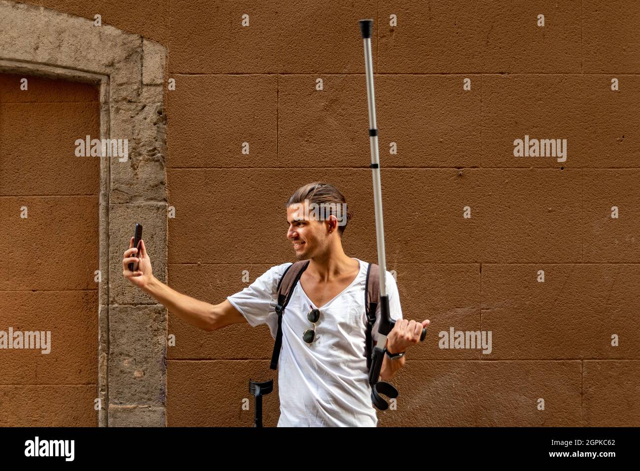 Giovane uomo con stampelle che prende un selfie con il suo cellulare per strada Foto Stock