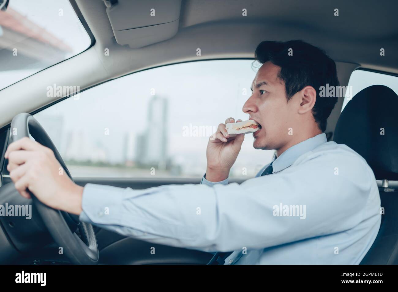 L'uomo d'affari sta mangiando un panino mentre guida su strada, e non è senza cintura di sicurezza. Foto Stock
