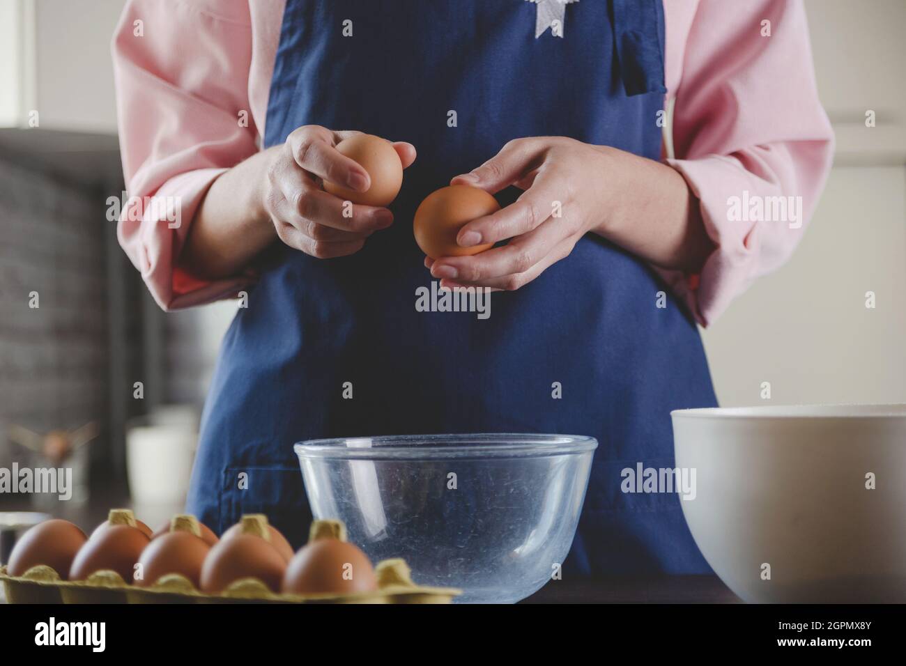 Donna panettiere in un grembiule blu rompe le uova per la cottura. Home cucina accogliente estetica. Foto Stock