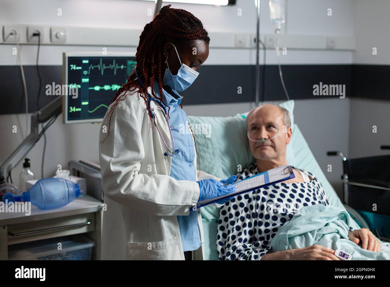 Medico afroamericano che indossa maschera chirurgica, in sala d'ospedale durante la consultazione di malato malato uomo anziano, respirando con l'aiuto di tubo di prova e fleboclisi iv attaccato. Foto Stock