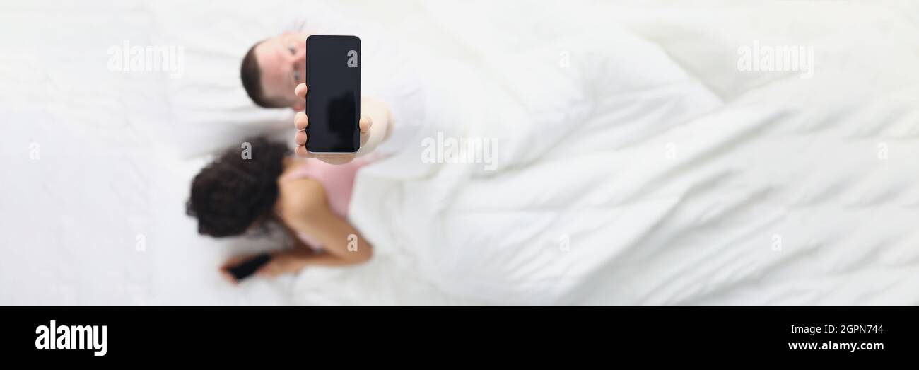 Donna che si allontana nasconde lo schermo dello smartphone dal marito mentre si trova a letto, marito la guarda utilizzando l'applicazione dal suo smartphone Foto Stock