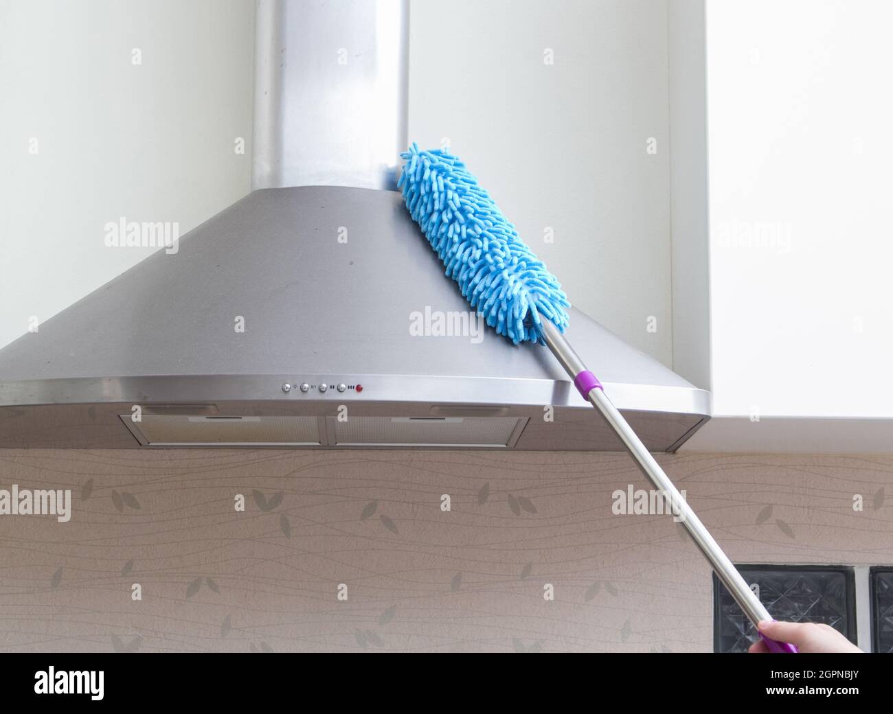 le mani della donna che tengono la piuma blu dello spolverino per pulire il cofano del fornello nella cucina Foto Stock