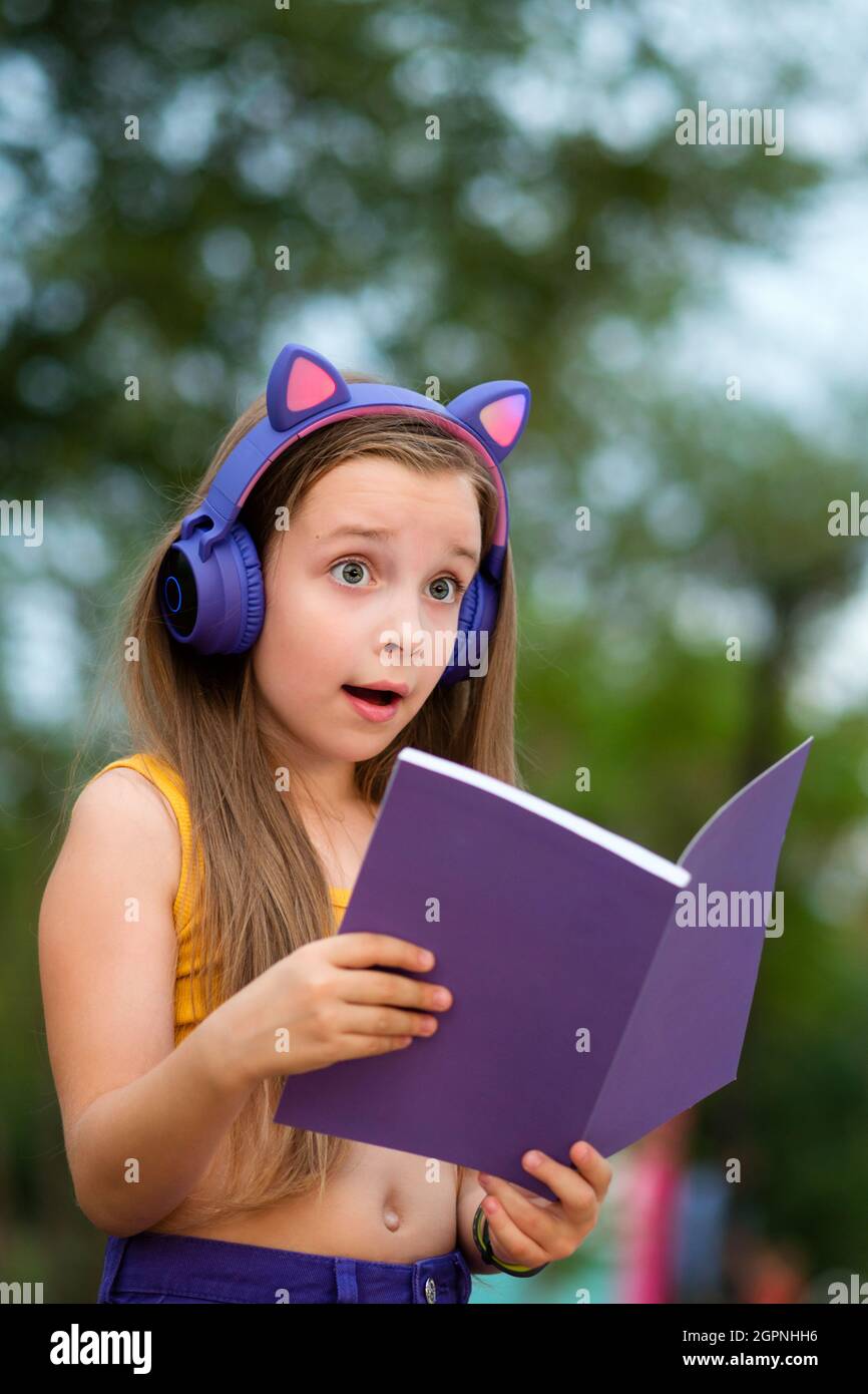 Sorpresa ragazza della scuola primaria che tiene un notebook aperto. Ragazza della scuola con libro su panca. I bambini con cuffie wireless indossano abiti alla moda, che si rilassano nel parco Foto Stock
