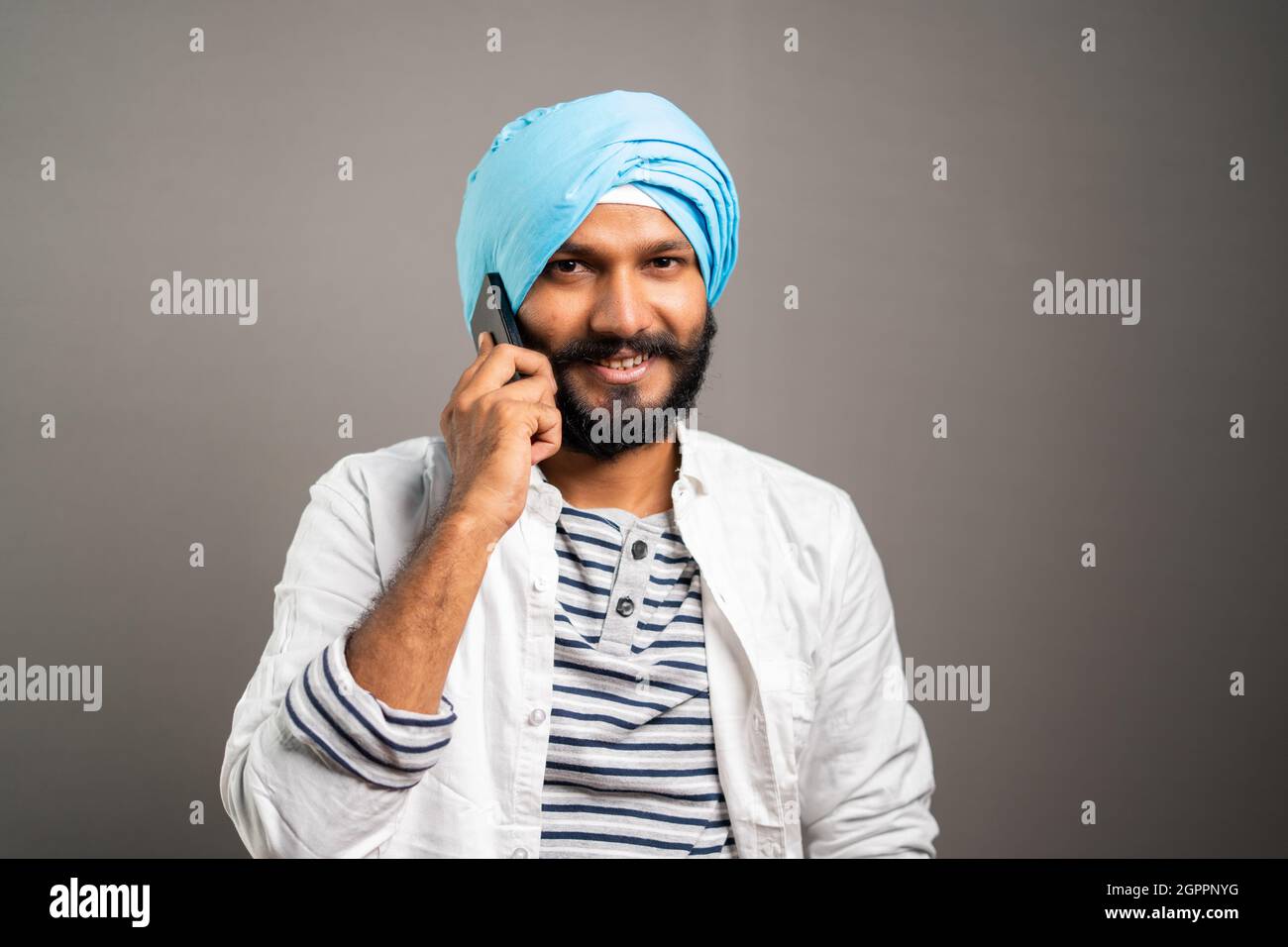 Felice sorridente indiano Sikh uomo occupato parlare sul telefono cellulare sullo sfondo studio guardando la fotocamera - concetto di comunicazione, rete e positivo Foto Stock