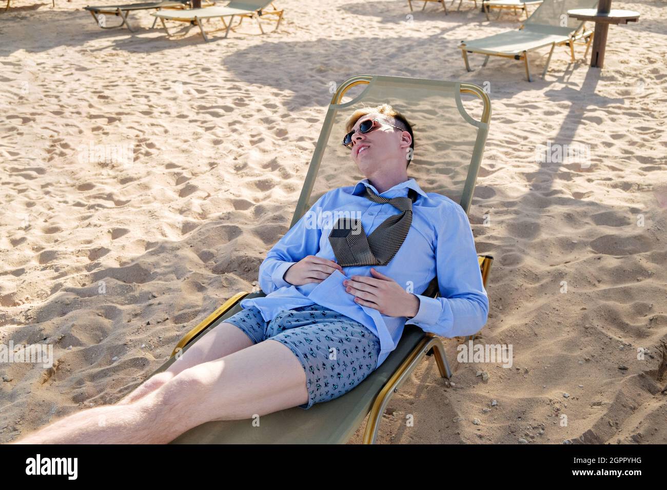 Giovane uomo d'affari stanco si trova su una chaise longue in una camicia con cravatta e boxer shorts. Riposati in mare, riavvia. Passaggio a una marcia inferiore Foto Stock