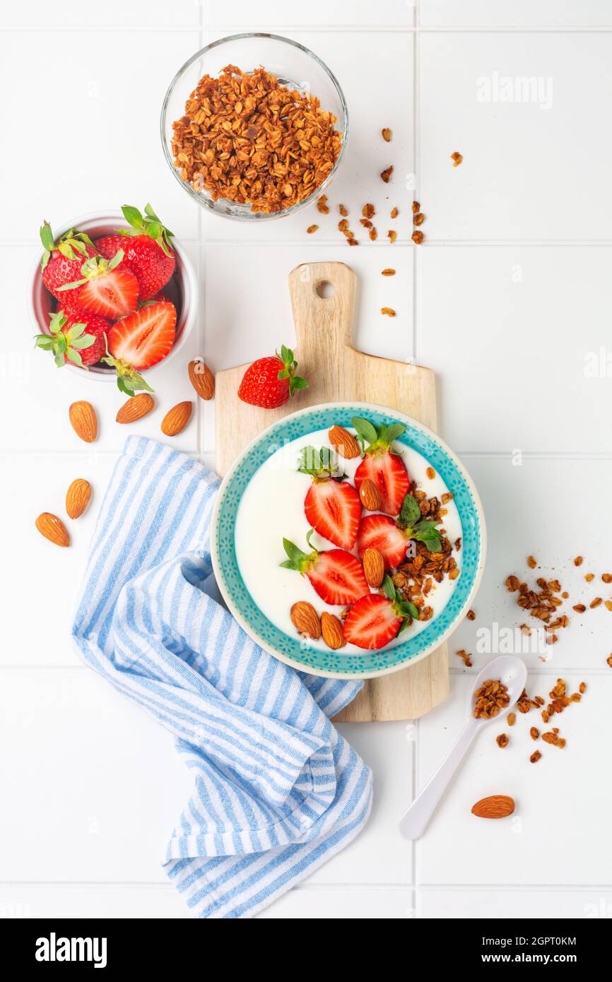 Yogurt greco in ciotola bianca con ingredienti per preparare la colazione a base di muesli e fragole fresche su tavola bianca. Vista dall'alto Foto Stock