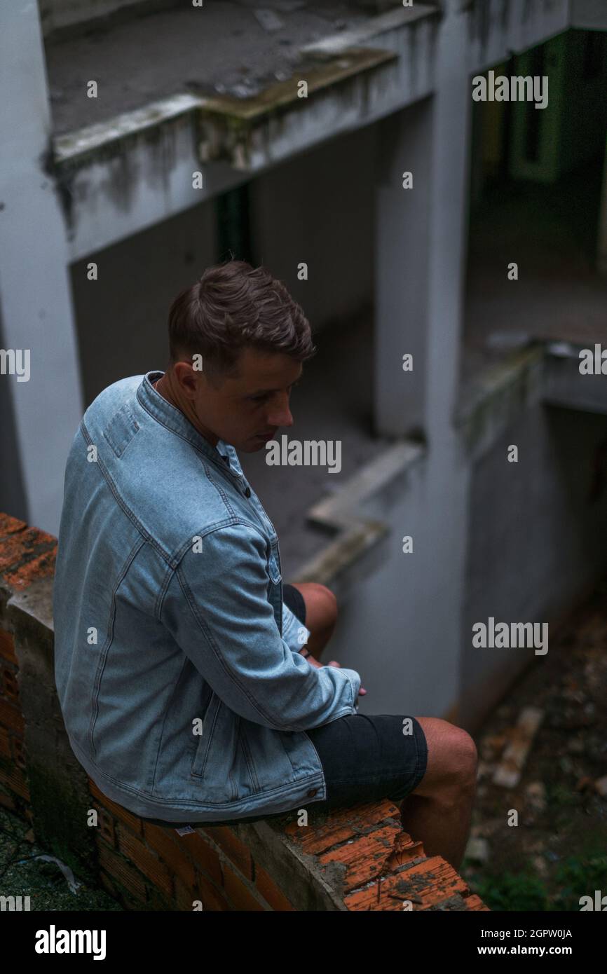 Ritratto di giovane bel caucasico in giacca denim, seduto sul cantiere abbandonato e guardando verso il basso Foto Stock