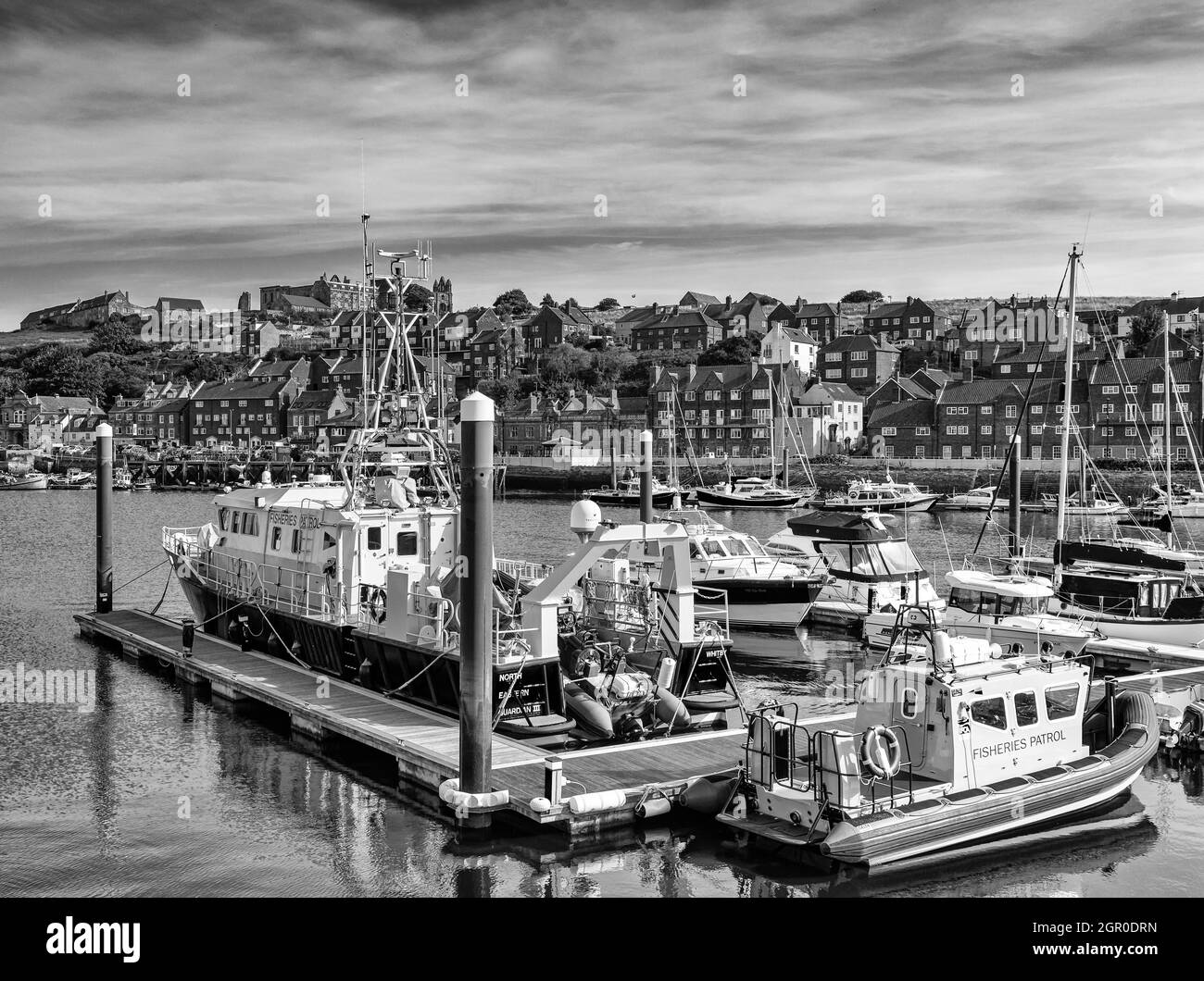 Una barca di pattuglia di pesca è ormeggiata in un porto. Edifici in mattoni rossi fiammeggiano il molo opposto e un cielo pieno di nuvole è sopra. Foto Stock