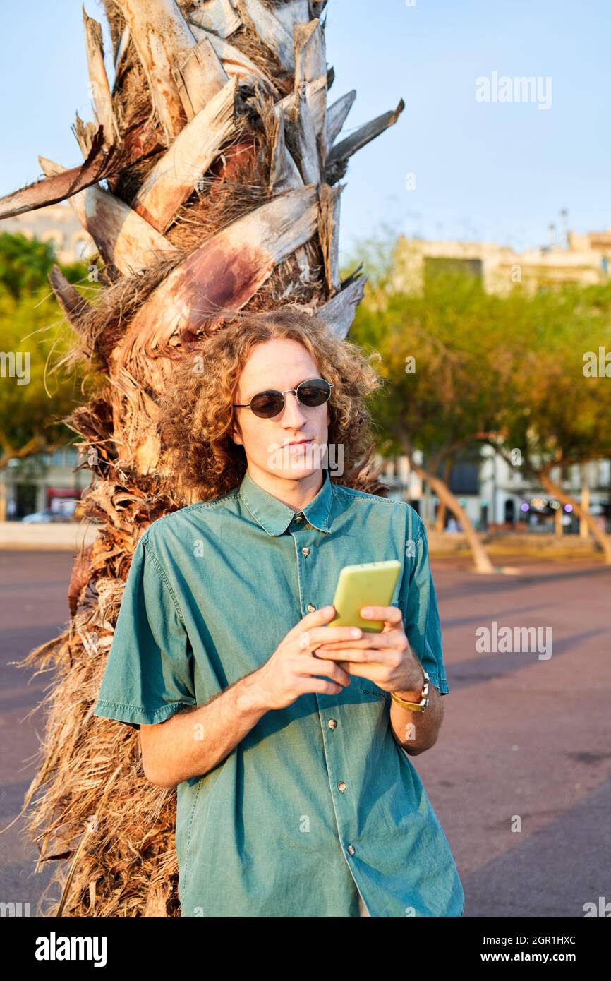 Giovane hipster ragazzo con capelli ricci e occhiali da sole è messaggi sul  telefono. È appoggiato su una palma Foto stock - Alamy