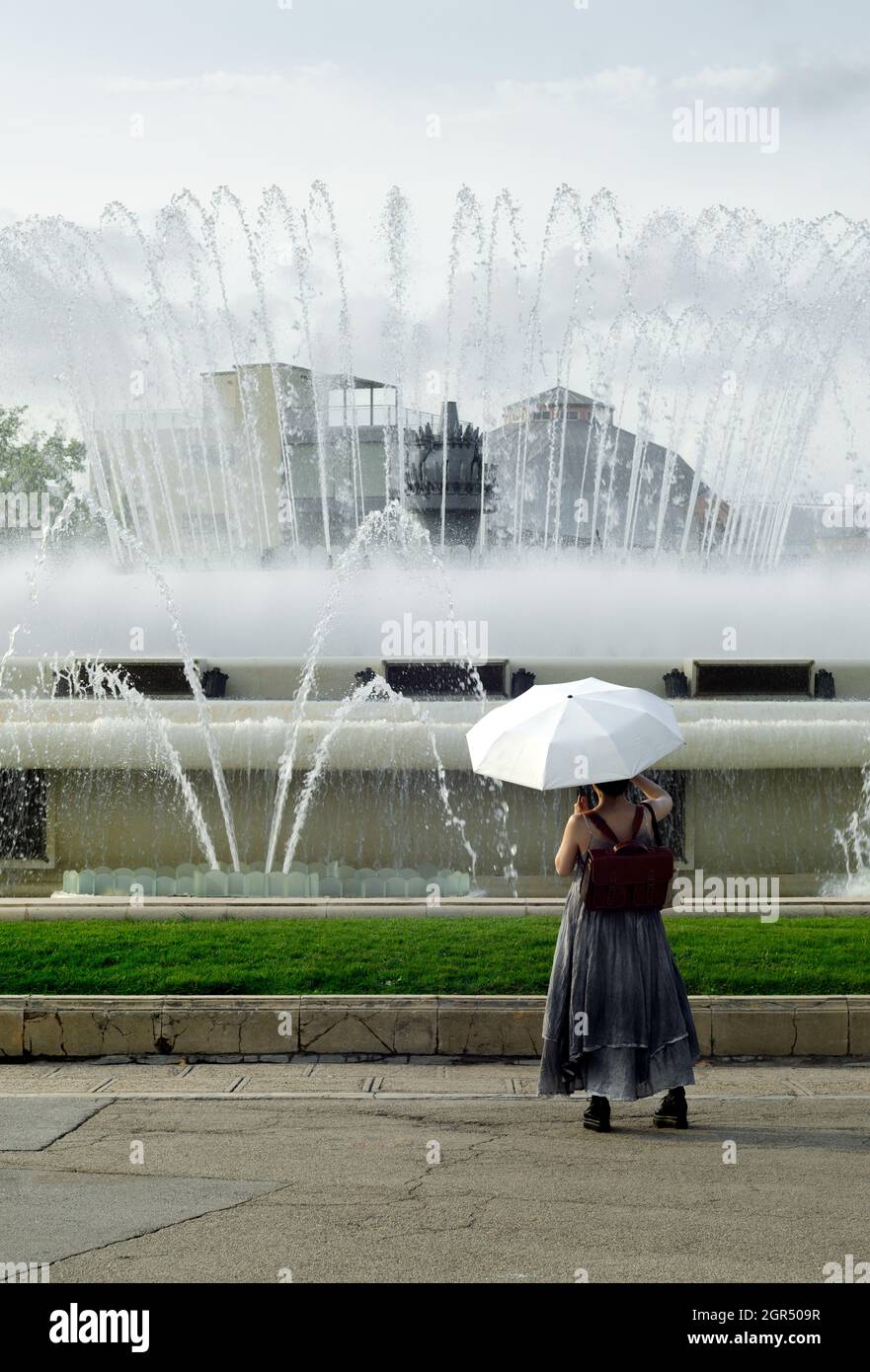 torna di giovane donna turistica che tiene un ombrello bianco dal sole guardando la magica fontana di barcellona in spagna. Donna che fa una foto di Foto Stock