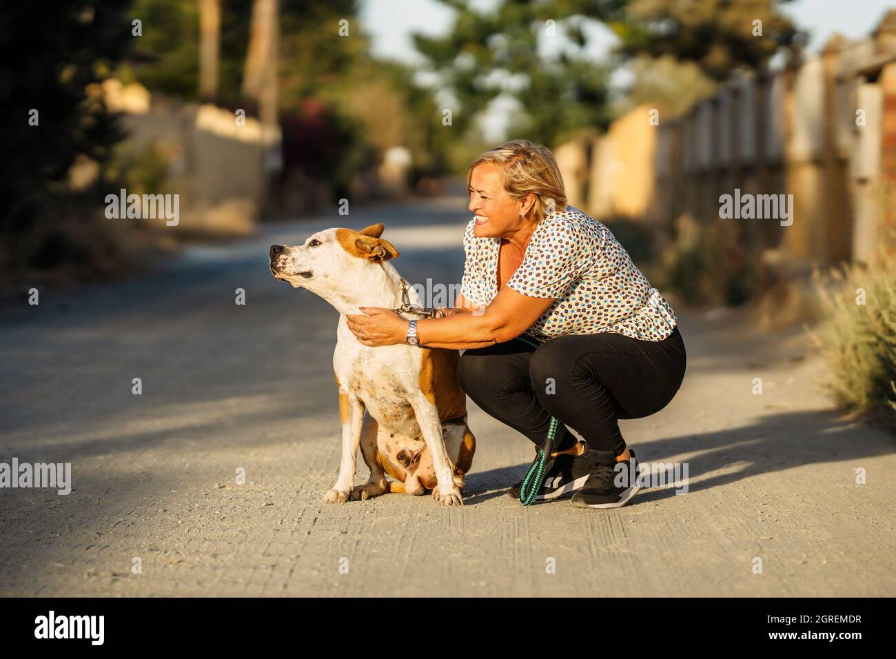 Donna accovacciata in una strada sterrata accanto ad un cane che si siede distratto Foto Stock
