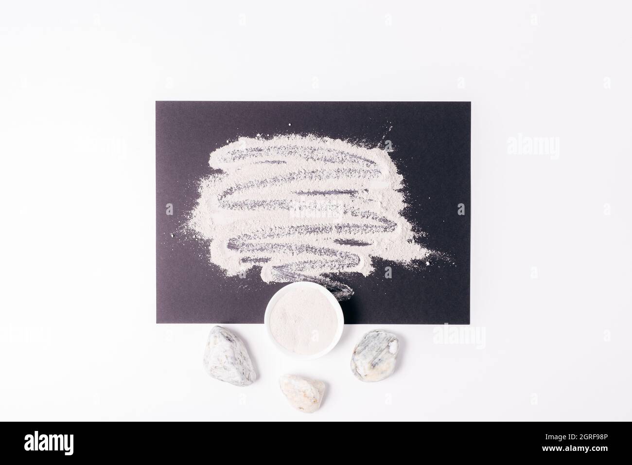 Polvere cosmetica organica di argilla sparsa su tavoletta nera accanto a pietre e ciotola, composizione di posa piatta Foto Stock