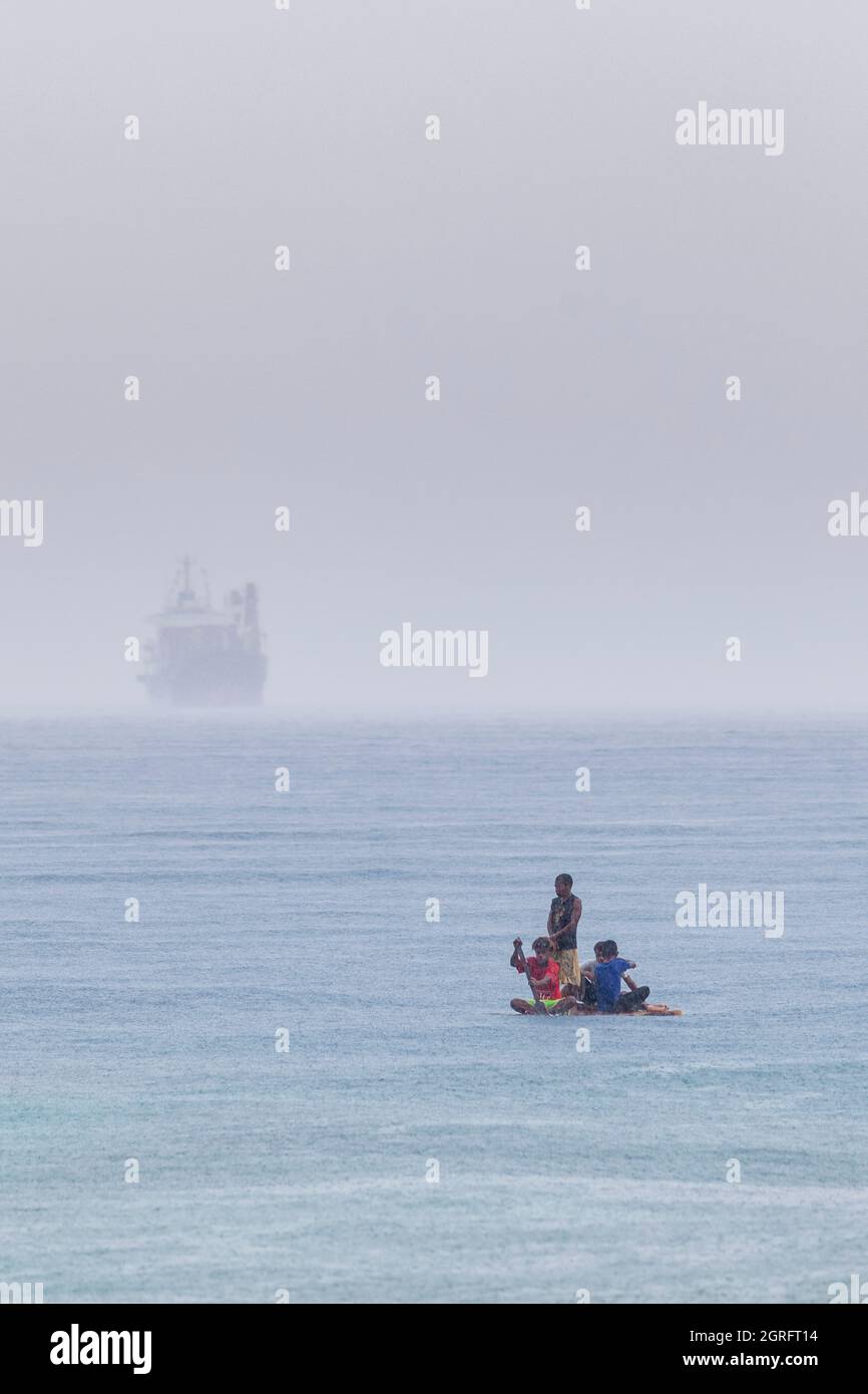 Indonesia, Papua, isola di Biak, bambini piccoli che giocano con una zattera sul mare e la silhouette di una nave container appare all'orizzonte Foto Stock