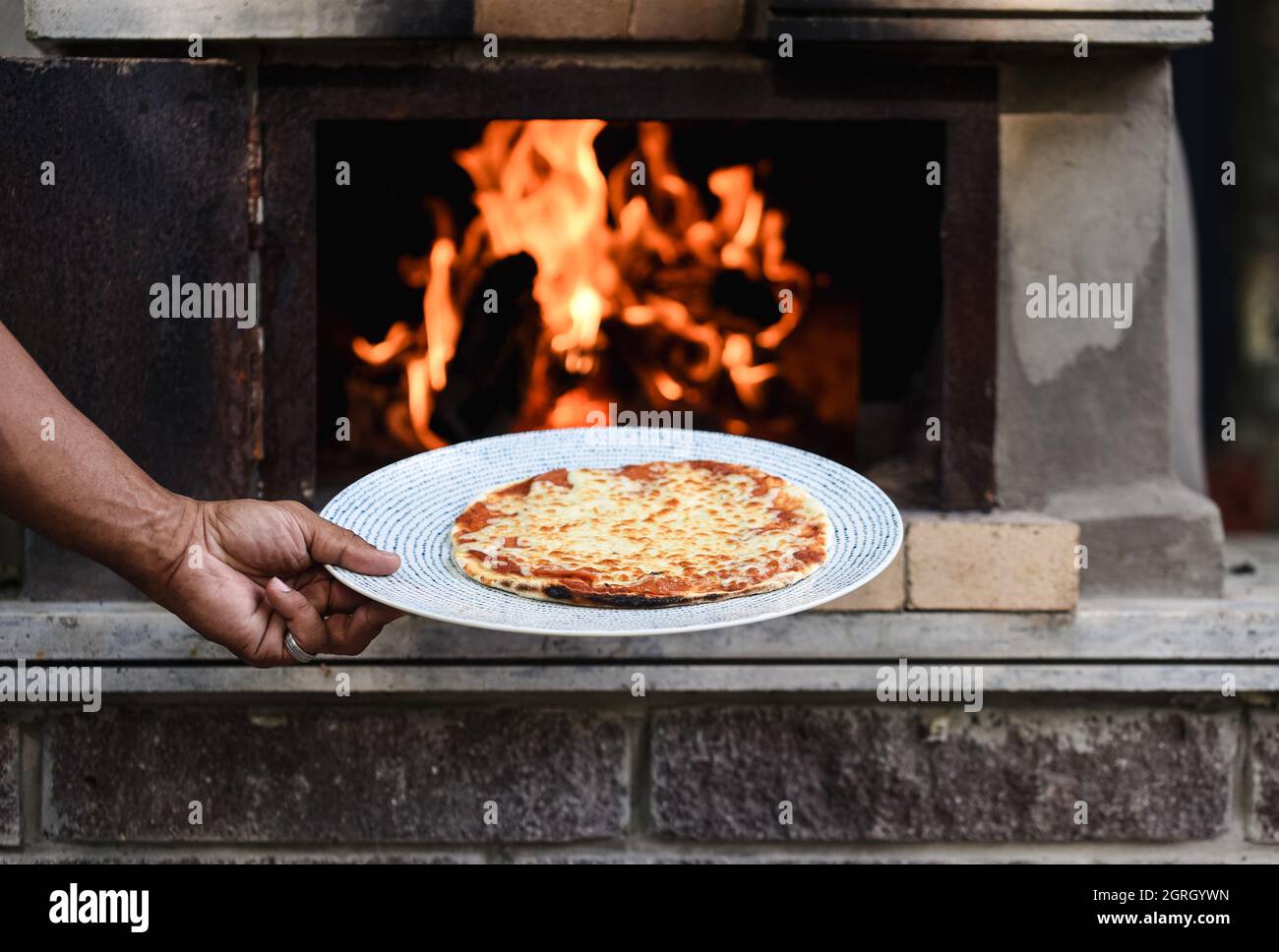 Braccio che tiene una pizza appena sfornata davanti ad un forno a legna per pizza. Foto Stock