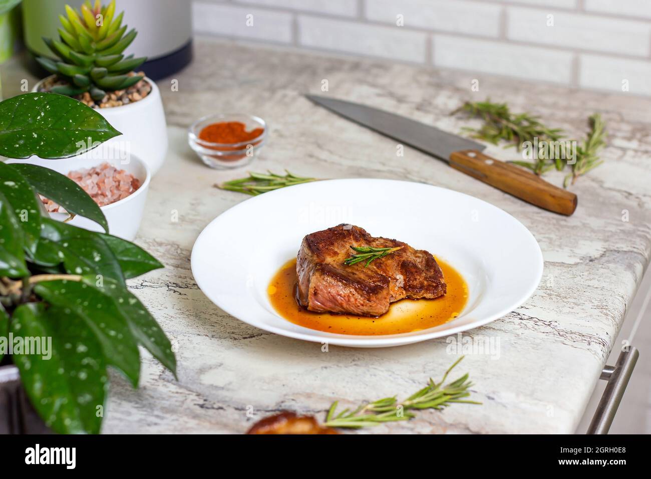 Bistecca di manzo arrosto preparata con spezie e rosmarino su piatto rotondo bianco sullo sfondo del tavolo da cucina. Foto Stock